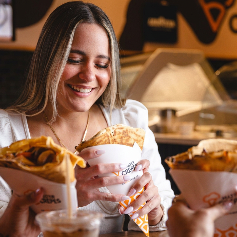 Smiling woman holds a filled crepe at a warmly lit restaurant table, with two other people holding crepes nearby.