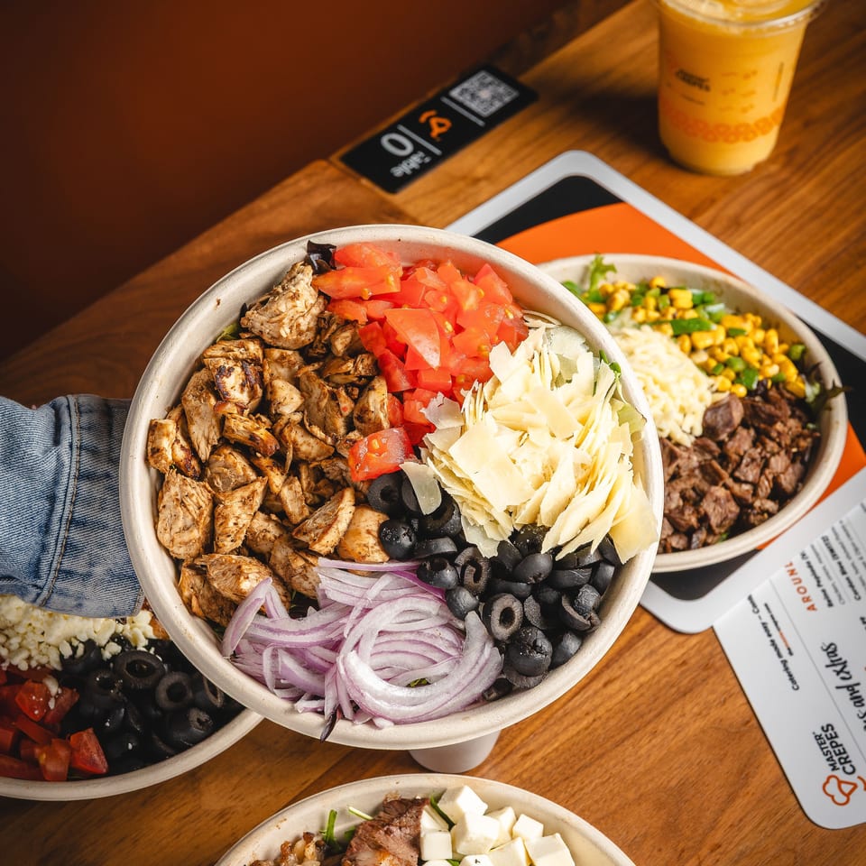 Greek-style grain bowls topped with grilled chicken, steak, tomatoes, olives, onions, cheese, and a golden iced drink on a wooden table.